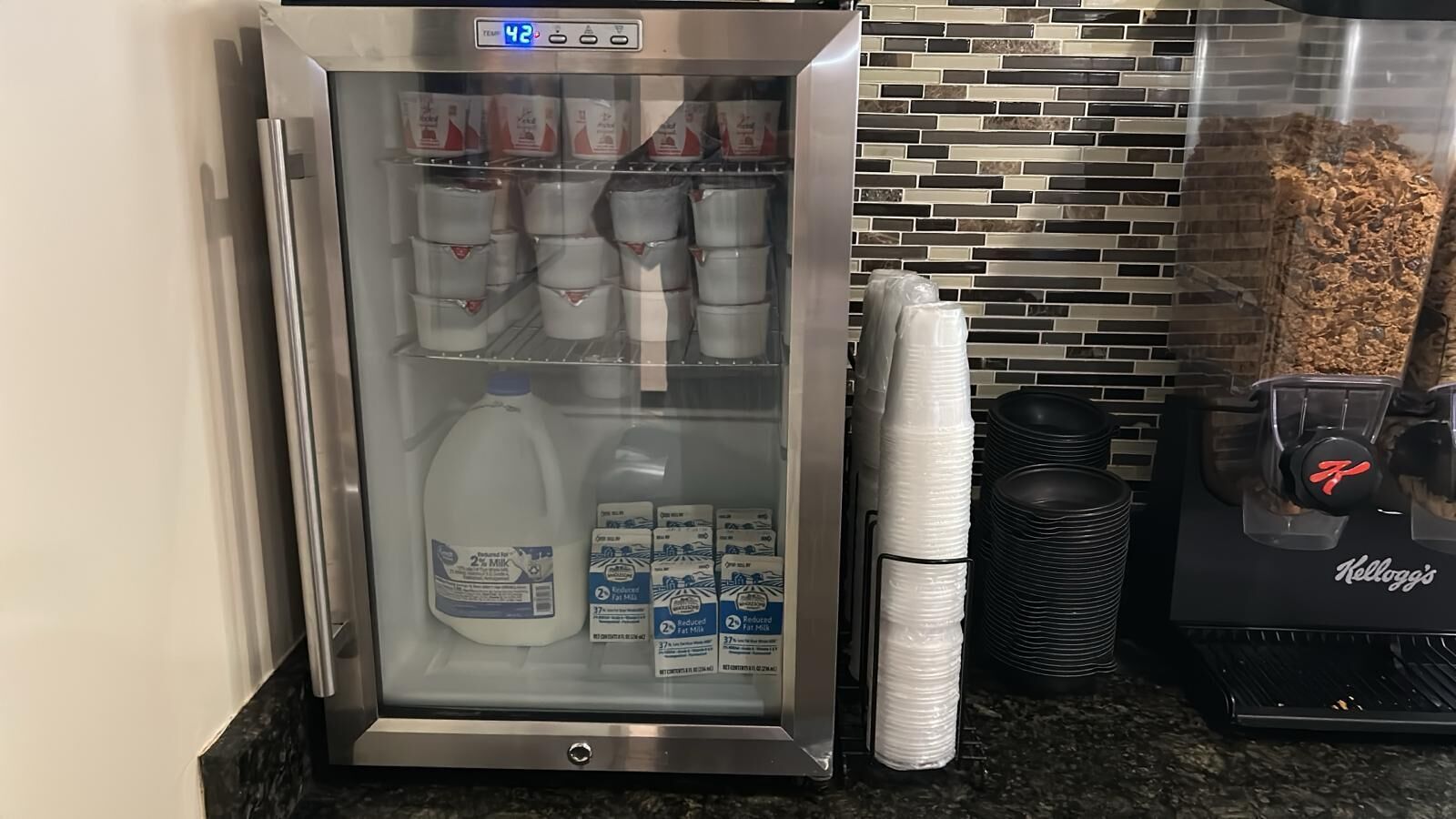 A refrigerator stocked with milk and juice cartons, a stack of plastic cups, and a Kellogg's branded coffee machine.