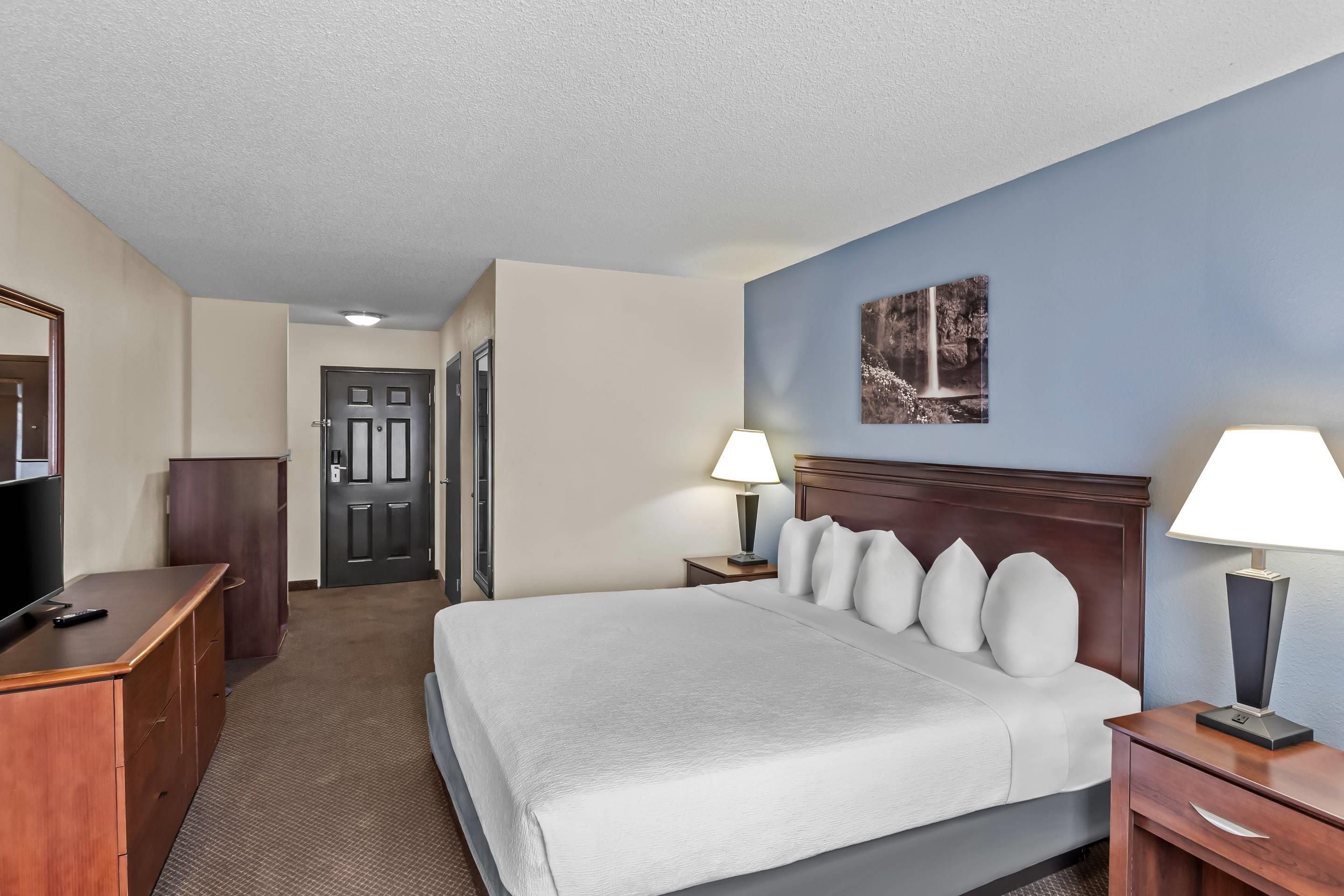 Quarto de hotel com cama, duas mesas de cabeceira com luminárias, televisão, guarda-roupa e porta que dá para outro espaço.