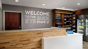 Zona de recepción de un hotel con un mostrador de madera, un cartel en la pared que dice "WELCOME TO THE BUCK EYE STATE" y un minibar con diversos artículos.