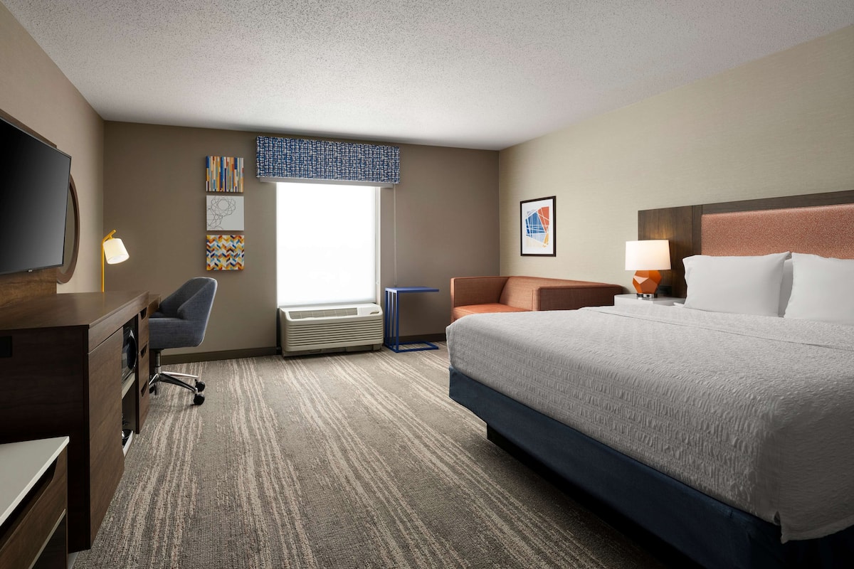 A hotel room with a large bed, a desk with a chair, a TV, a window with blinds, and artwork on the walls.