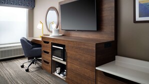 Habitación de hotel con un escritorio de madera, un televisor de pantalla plana, un espejo redondo, una lámpara de escritorio y una cafetera.