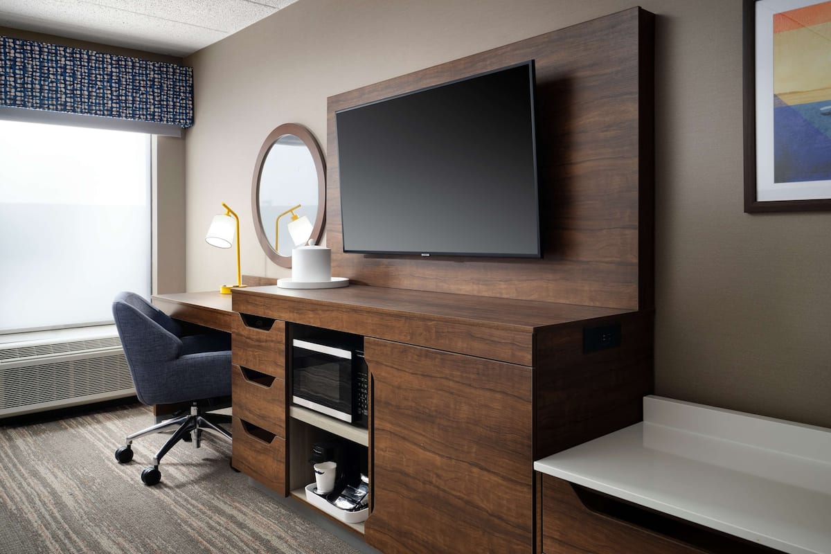 A hotel room with a wooden desk, a flat-screen TV, a round mirror, a desk lamp, and a coffee machine.