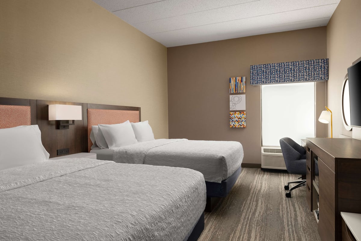 A hotel room with two large beds, a desk with a chair, a television, and artwork on the walls.