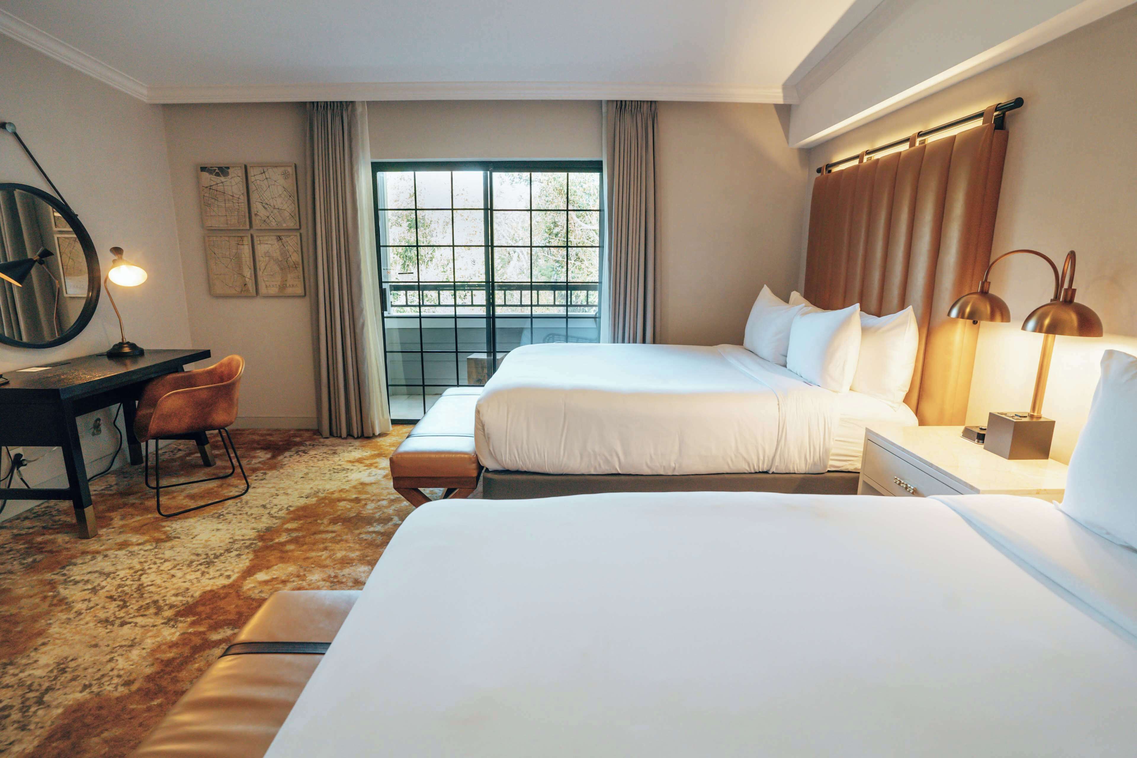 A hotel room with two beds, a desk, a chair, a large window with curtains, and a mirror.