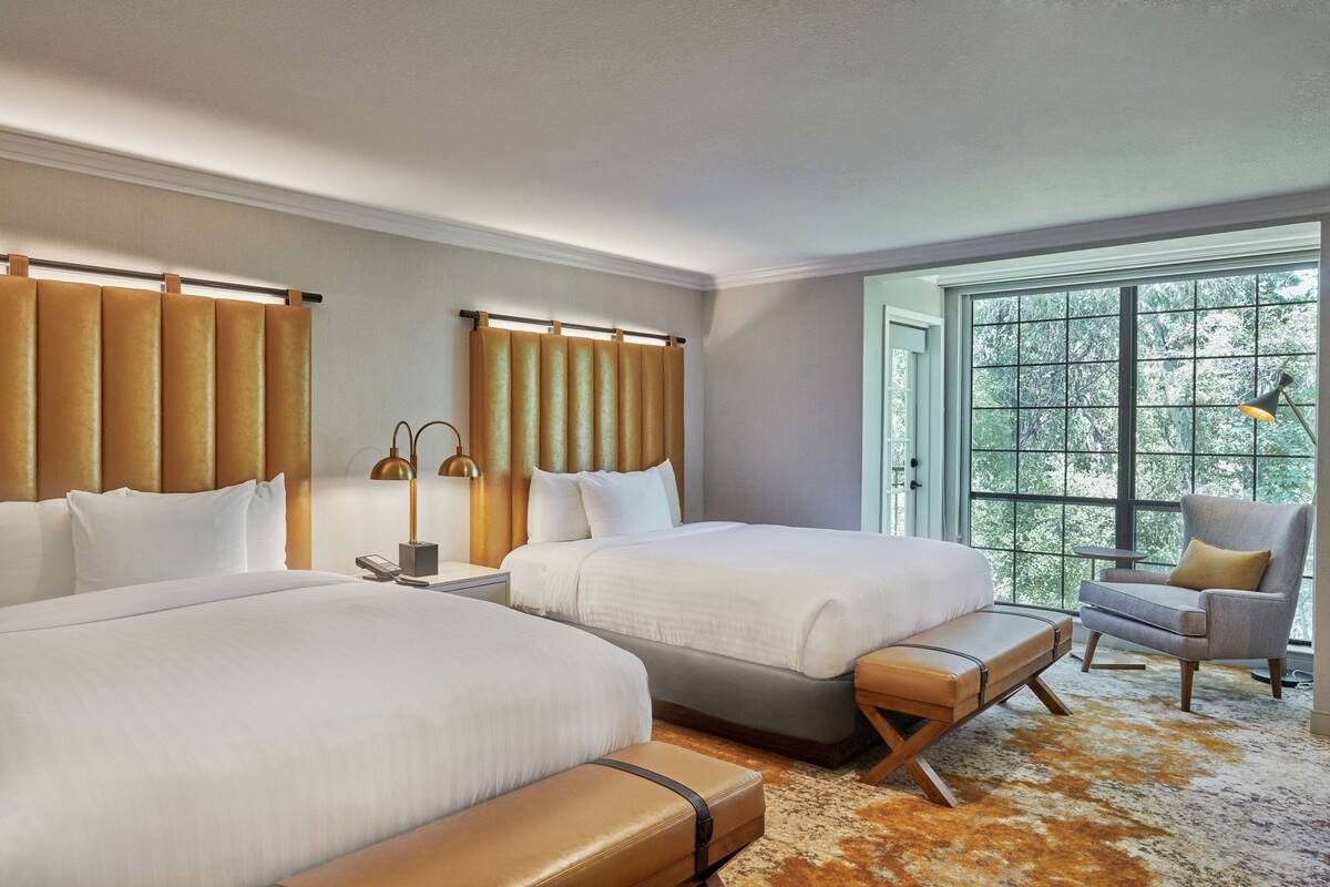 A hotel room with two beds, a large window with a view of trees, a chair, and a lamp.