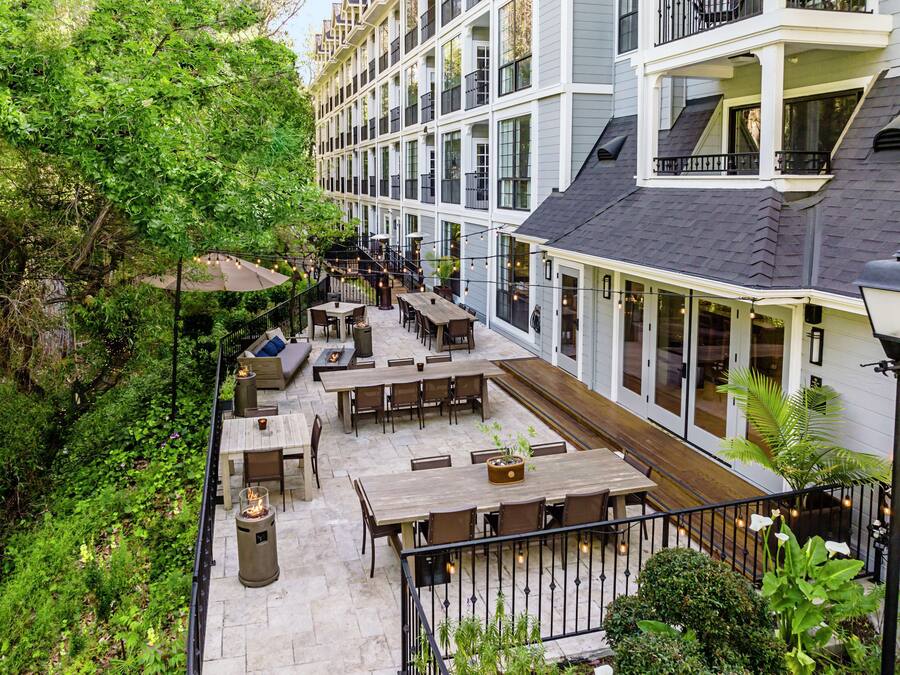 An outdoor patio with multiple tables and chairs, surrounded by lush greenery and a building with large windows.