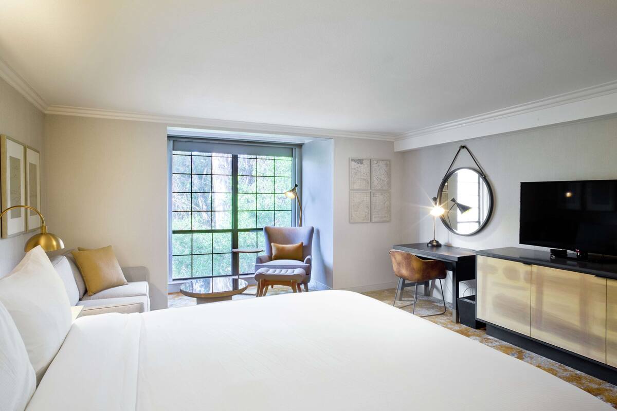 A hotel room with a large bed, a desk, a chair, a television, and a window with a view of trees.