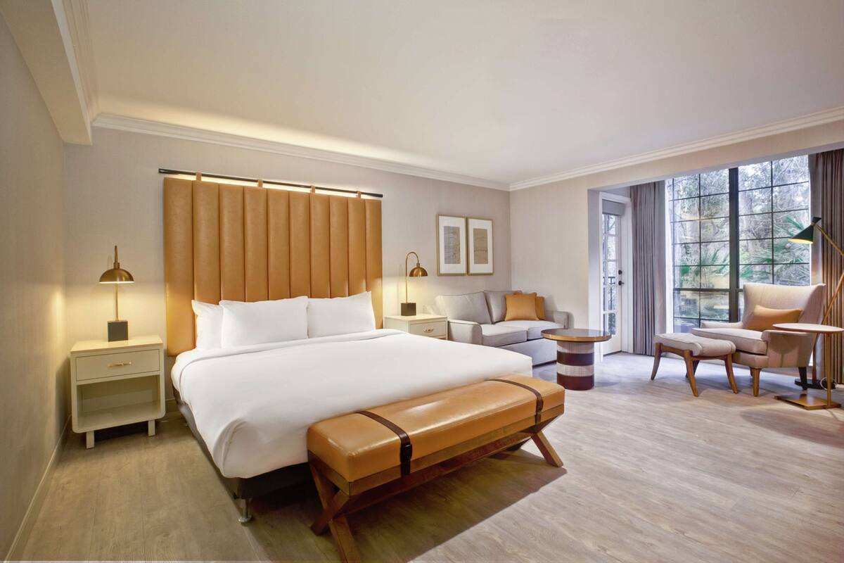 A modern hotel room with a large bed, a bench, a sofa, a desk, and a view of the outdoors.