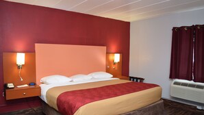 A hotel room with a large bed, two bedside lamps, a nightstand with a phone, and an air conditioning unit. - Econo Lodge Brockport (Brockport)