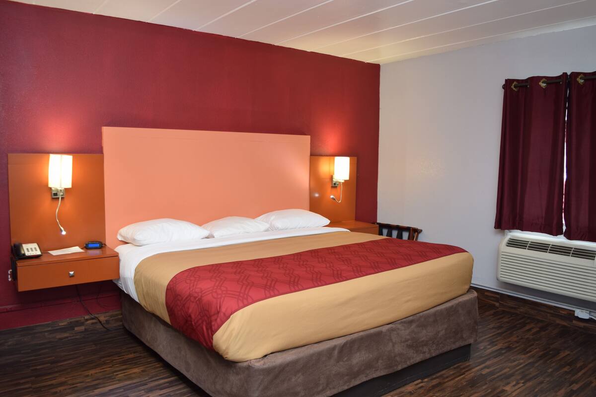 A hotel room with a large bed, two bedside lamps, a nightstand with a phone, and an air conditioning unit.
