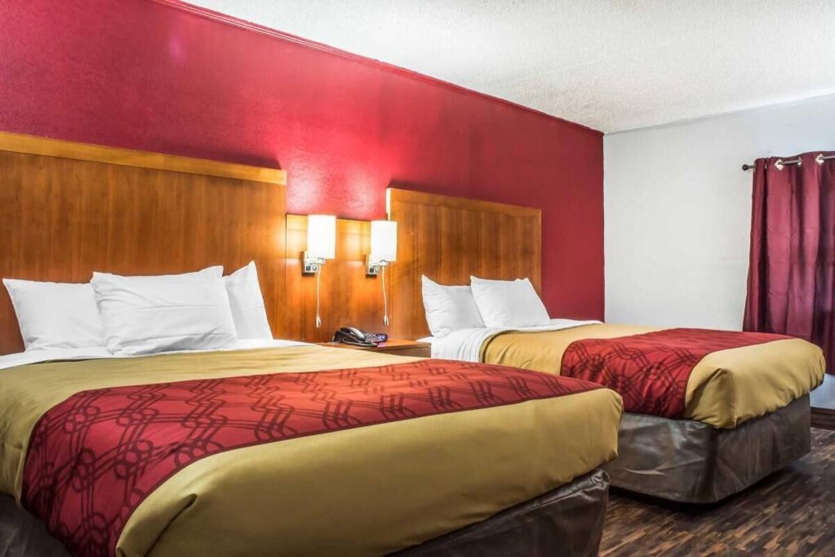 A hotel room with two beds, wooden headboards, wall-mounted lamps, and a red curtain.