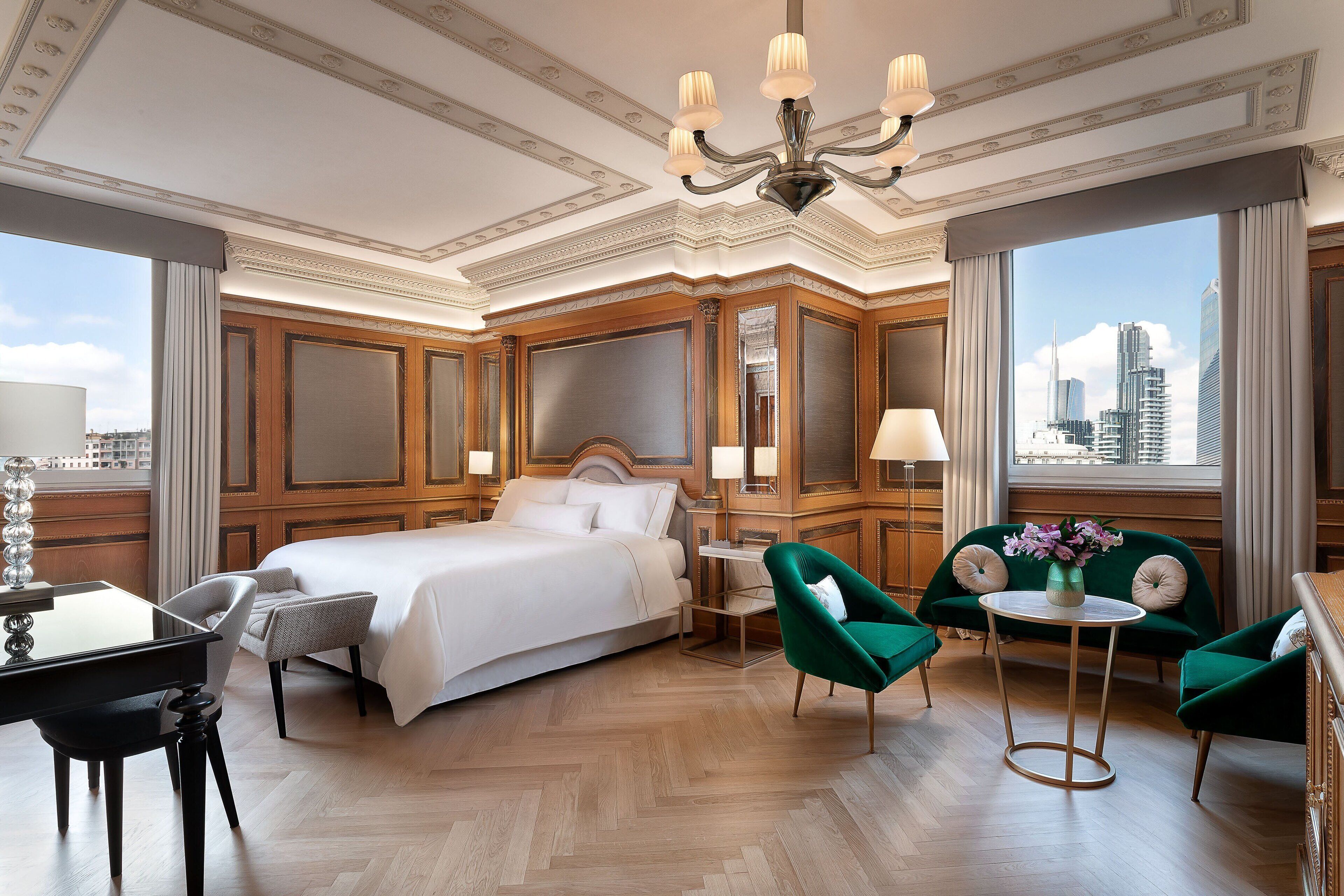 A hotel room with a large bed, a desk, two green armchairs, a round table, and a view of the city skyline.