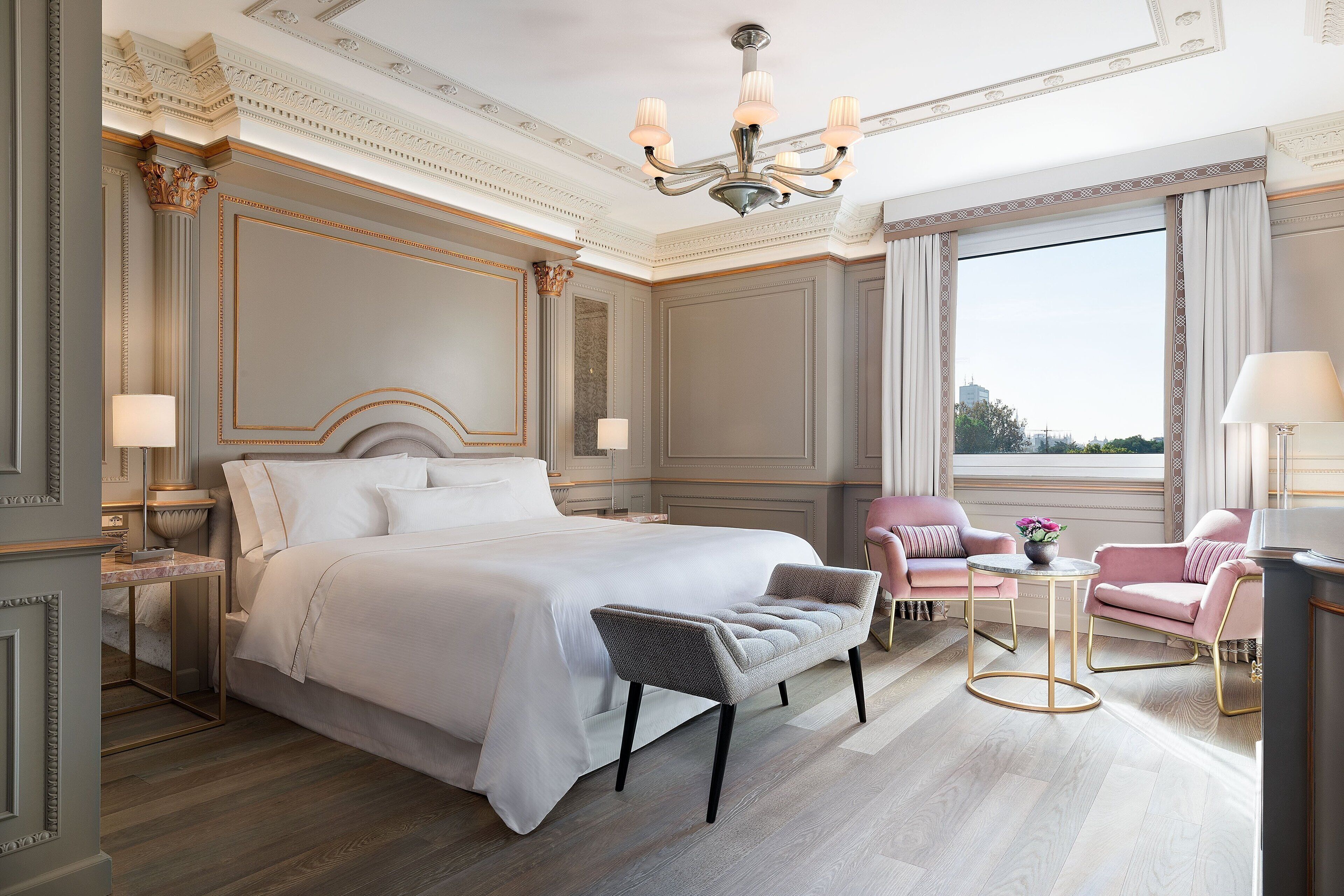 Un'ampia camera da letto con un letto grande, una chaise longue, un tavolino rotondo e vista sulla città attraverso ampie finestre.