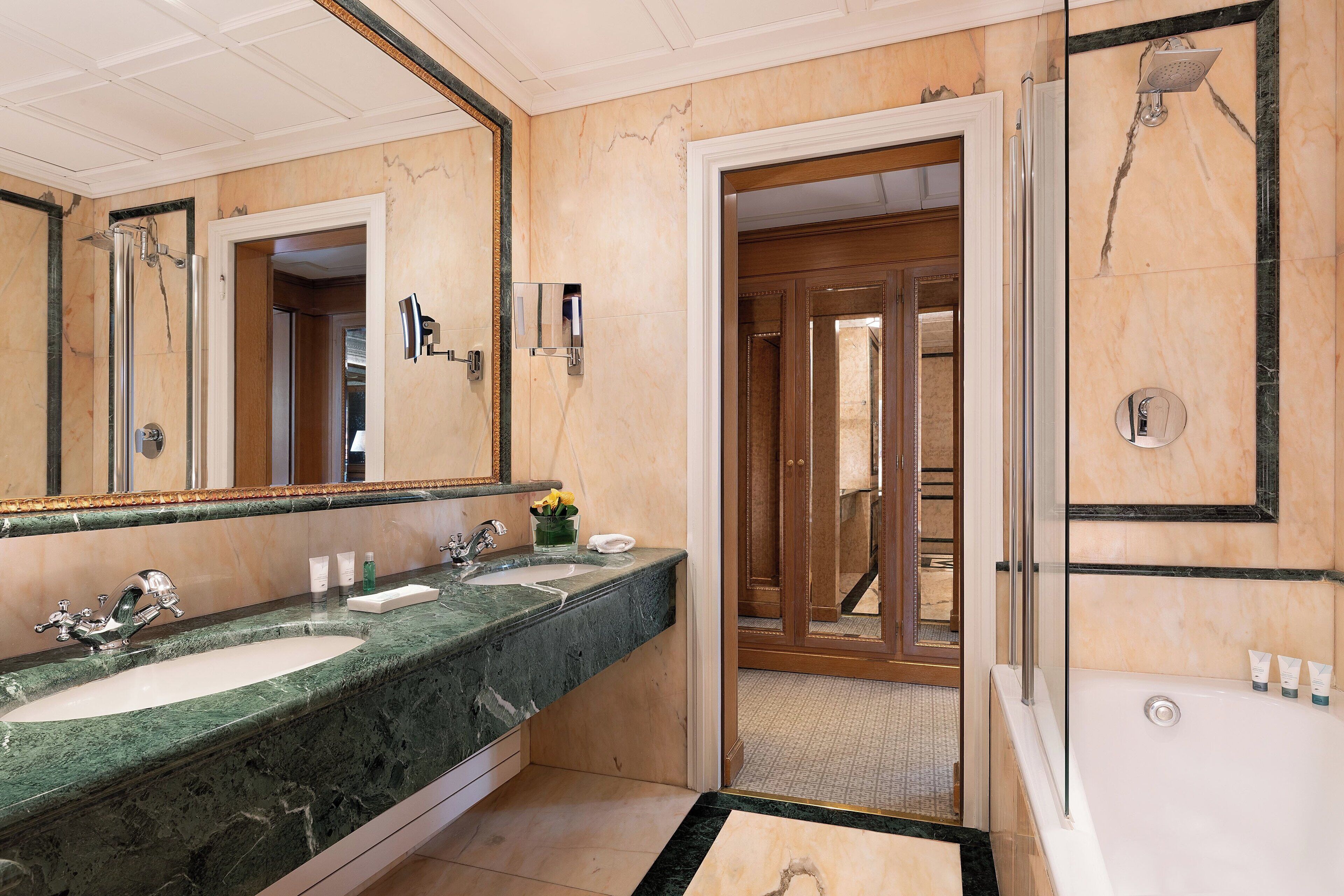 A bathroom with two sinks, a large mirror, a shower, and a door leading to another room.