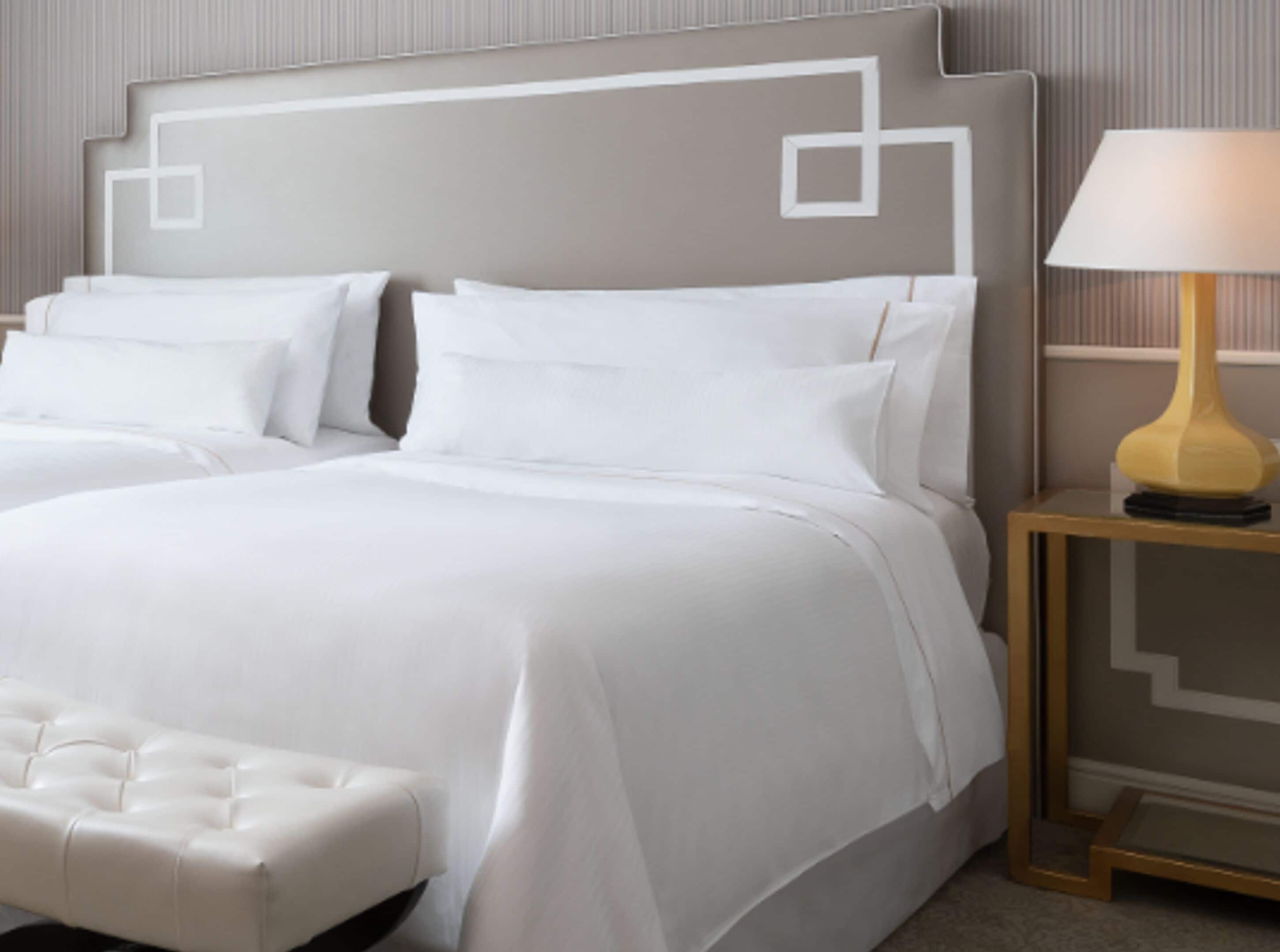 A neatly made bed with white linens and pillows, a tufted white bench, and a bedside table with a lamp.