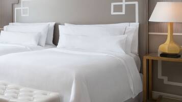 A neatly made bed with white linens and pillows, a tufted white bench, and a bedside table with a lamp.