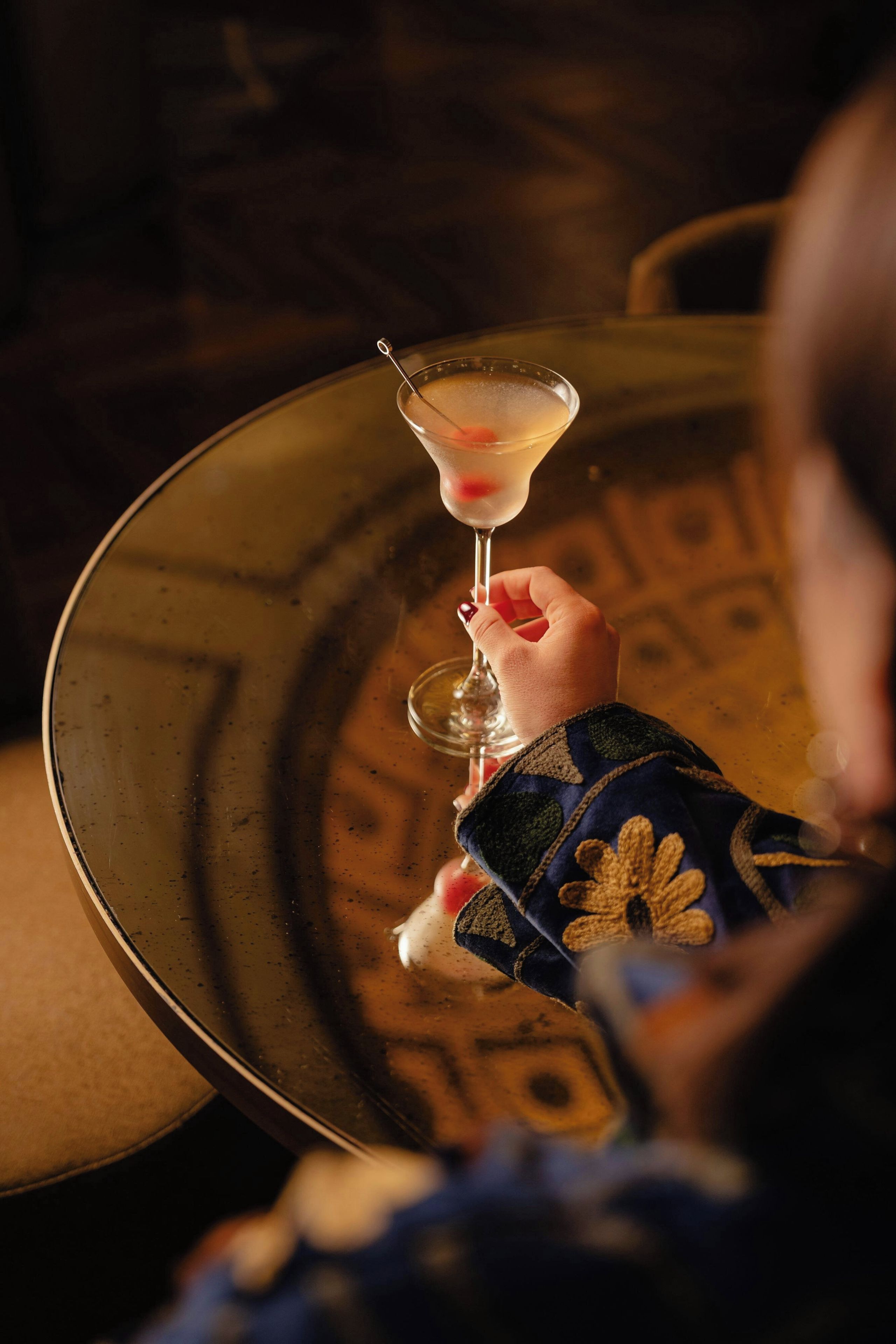A person holding a martini glass with a straw, with a reflective surface in the background.