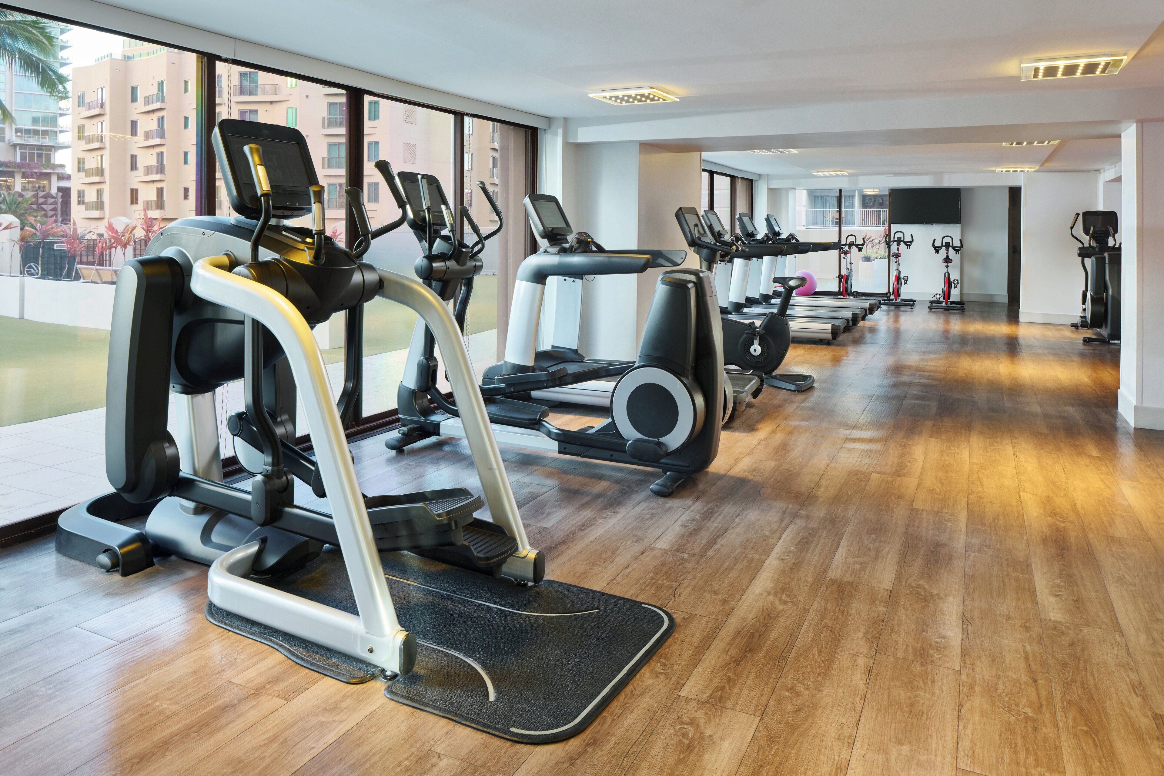 Una palestra con diverse macchine cardio, pavimenti in legno e ampie finestre che offrono vista su edifici e aree verdi.