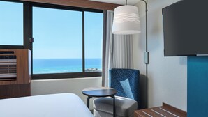 A hotel room with a large window offering a view of the ocean, a bed, a chair, a small table, and a flat-screen TV mounted on the wall. - Holiday Inn Express Waikiki by IHG (Honolulu)