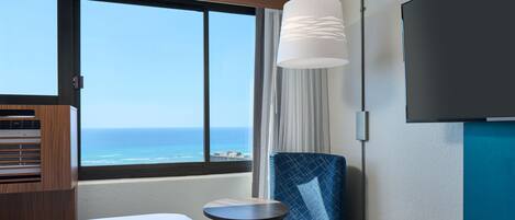 A hotel room with a large window, a bed, a chair, a small table, and a TV mounted on the wall.