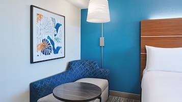 A hotel room with a blue accent wall, a framed abstract art piece, a round table, a blue upholstered bench, and a bed with white linens.