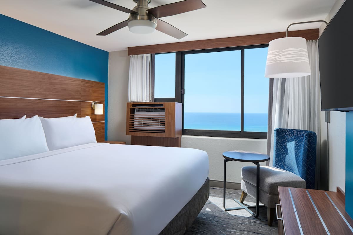 A hotel room with a large bed, a desk, a chair, a ceiling fan, a window with a view of the ocean, and a small table.