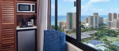 A compact room with a view of a cityscape and the ocean. It includes a microwave, a coffee machine, and a seating area with a view.