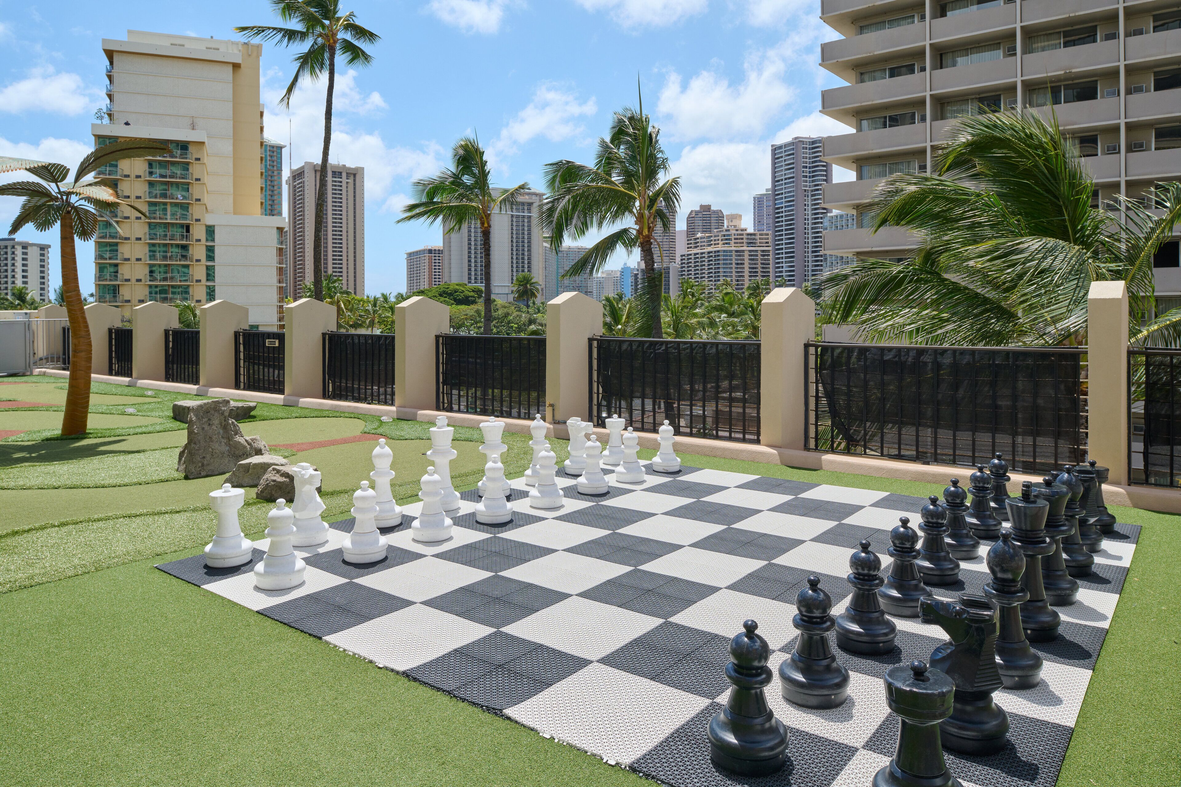 A large outdoor chessboard set up on artificial grass with a cityscape in the background.