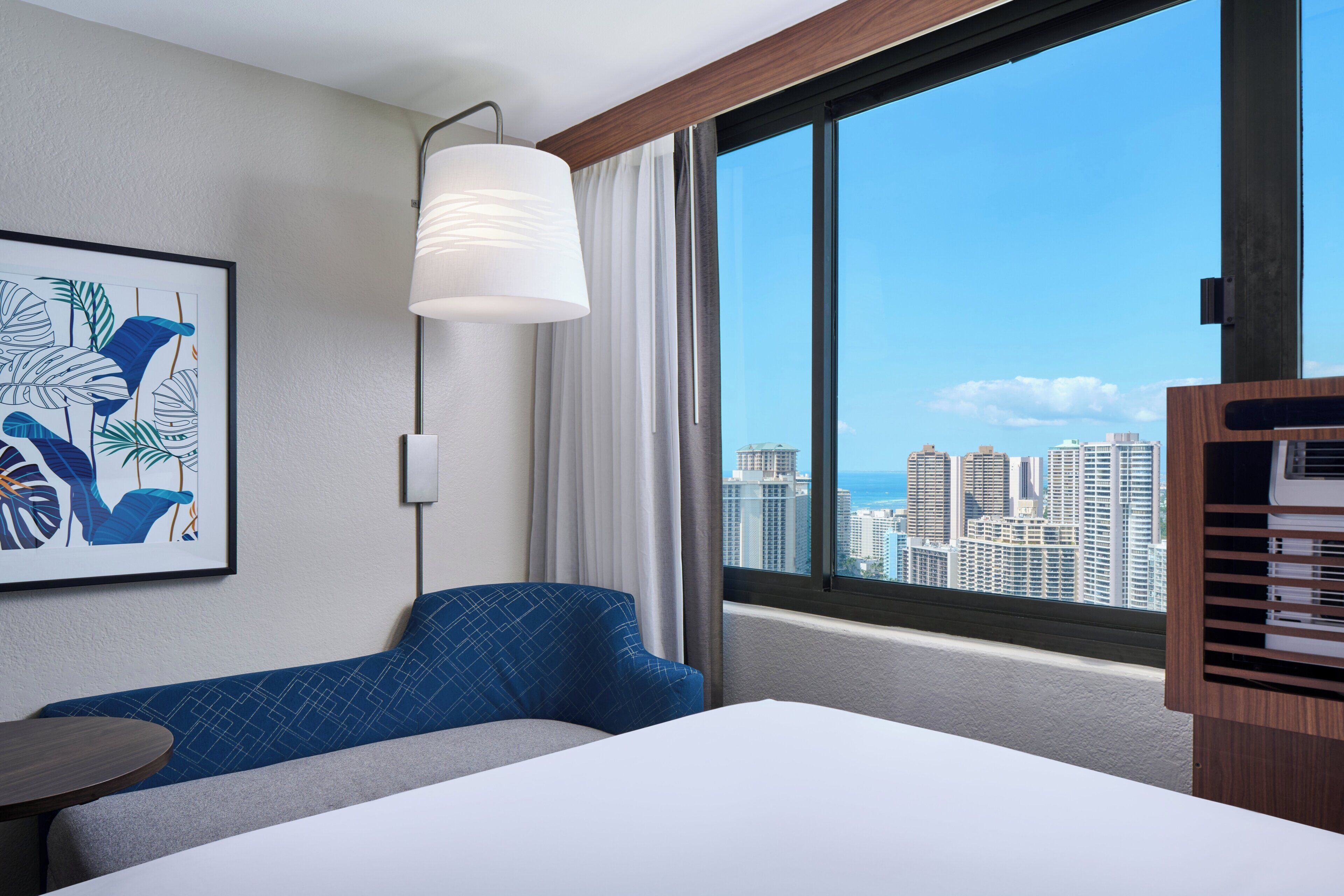 A hotel room with a bed, a blue sofa, a table, a large window with a city view, and a wall-mounted artwork.