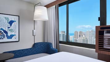 A hotel room with a bed, a blue sofa, a table, a large window with a city view, and a wall-mounted artwork.