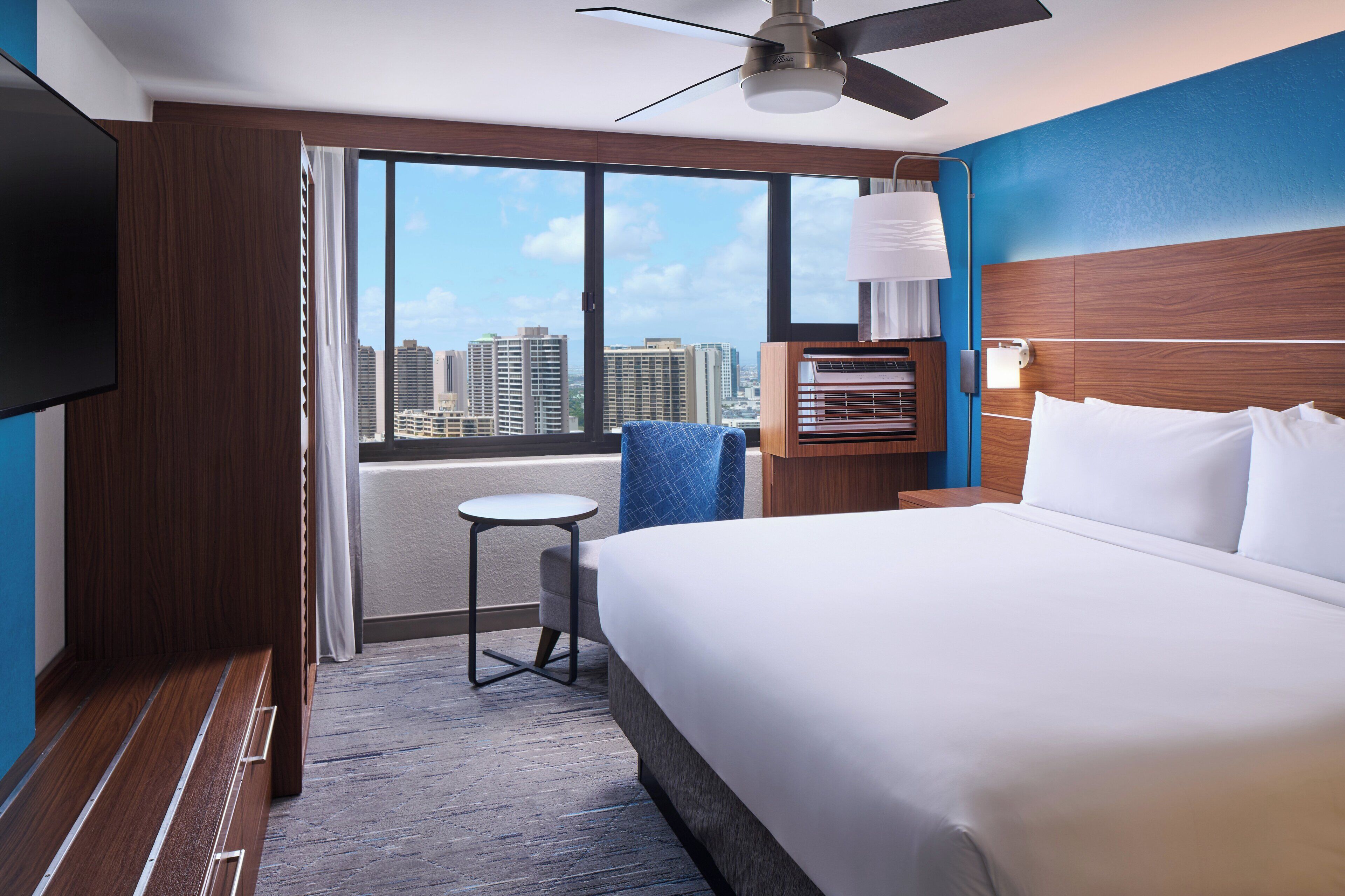 Habitación de hotel con una cama grande, televisión, ventilador de techo, una mesita y vistas a la silueta urbana a través de amplias ventanas.