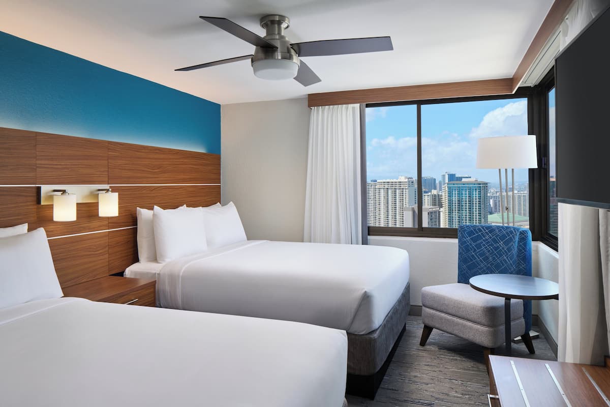 A hotel room with two beds, a ceiling fan, a city view, a small table, and a chair.