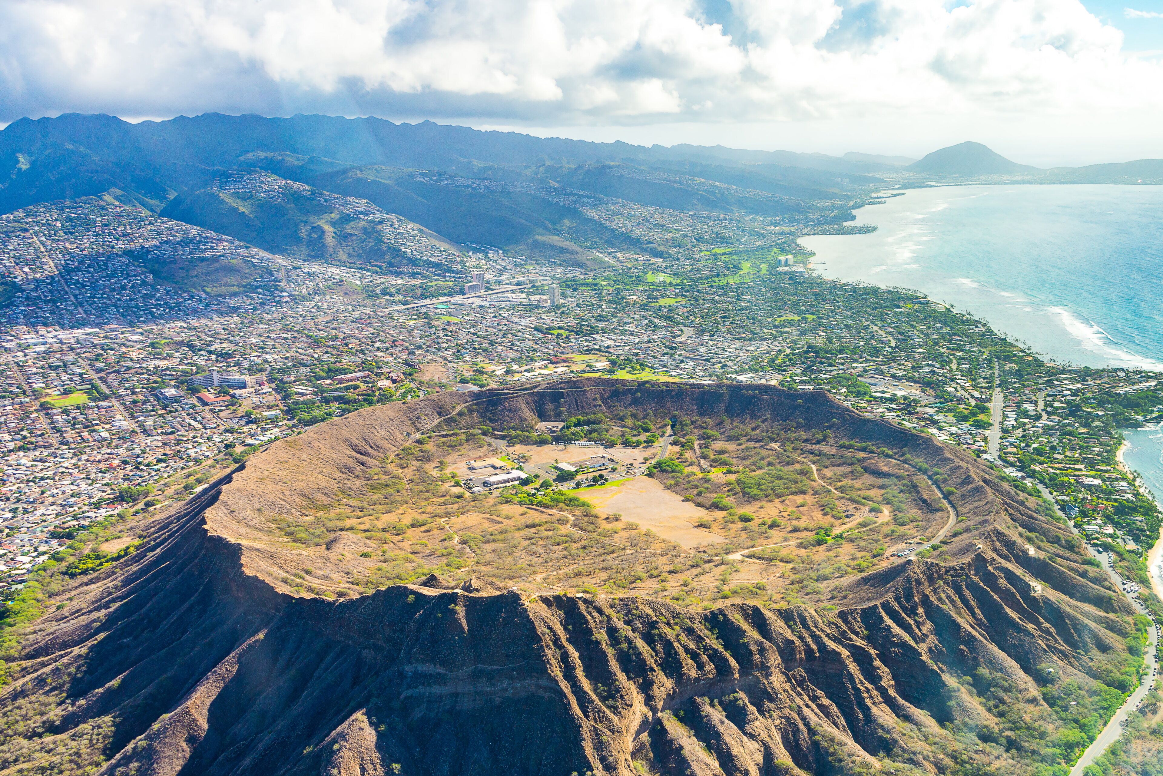 Vue aérienne d’une ville côtière, avec un cratère volcanique bien visible, entouré de zones résidentielles et bordé d’une plage.