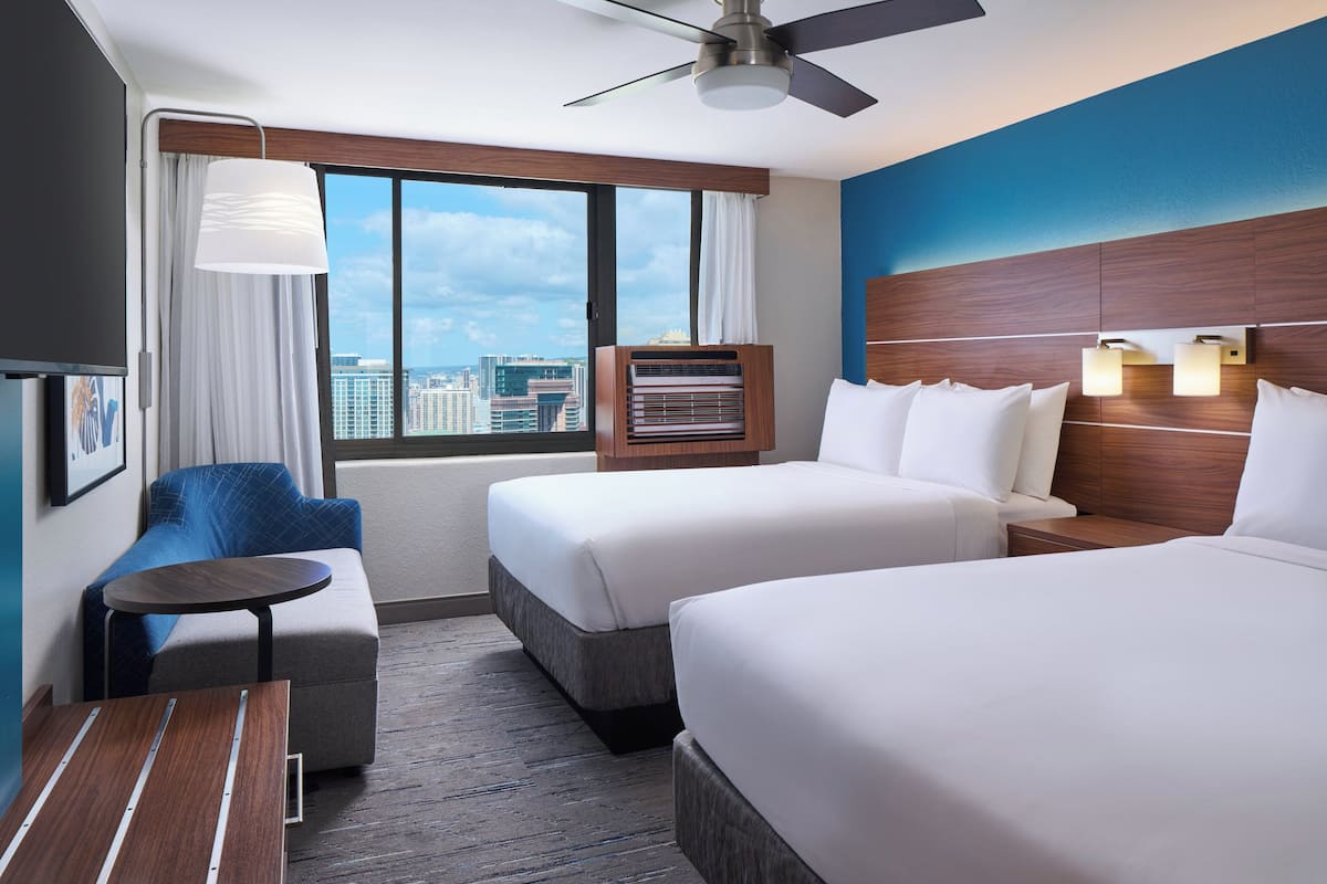 A hotel room with two beds, a sofa, a small table, a ceiling fan, and a large window with a city view.