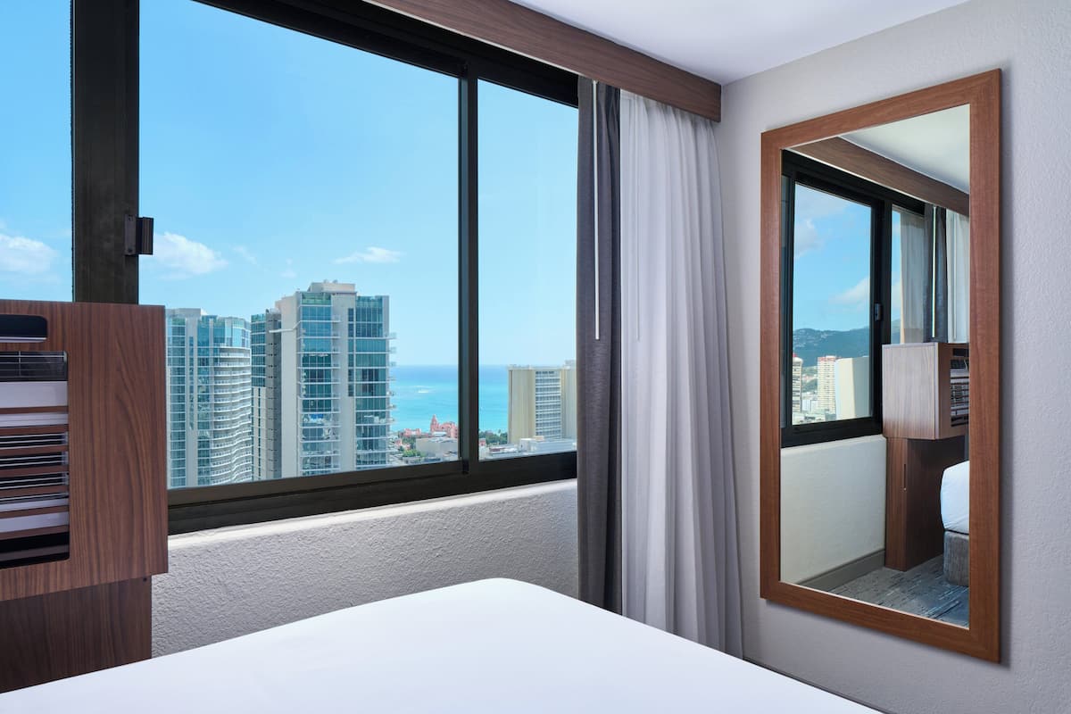 A hotel room with a large window, a bed, a mirror, and a wooden cabinet.