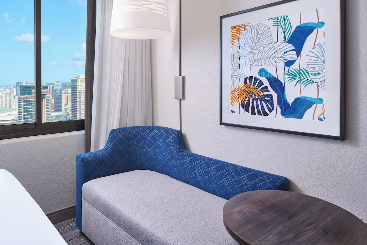 A modern hotel room with a large window, a blue patterned sofa, a small round table, and a framed abstract art piece on the wall.