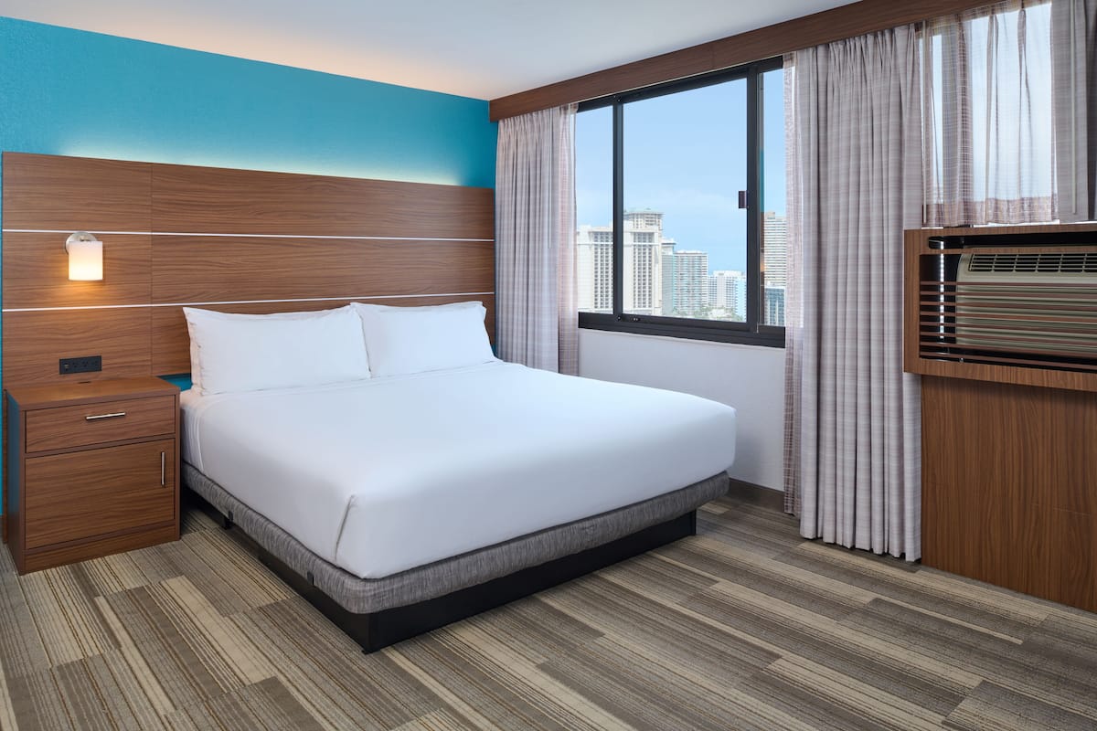 A hotel room with a large bed, wooden headboard, a nightstand, a bedside table, a window with curtains, and a view of the city skyline.