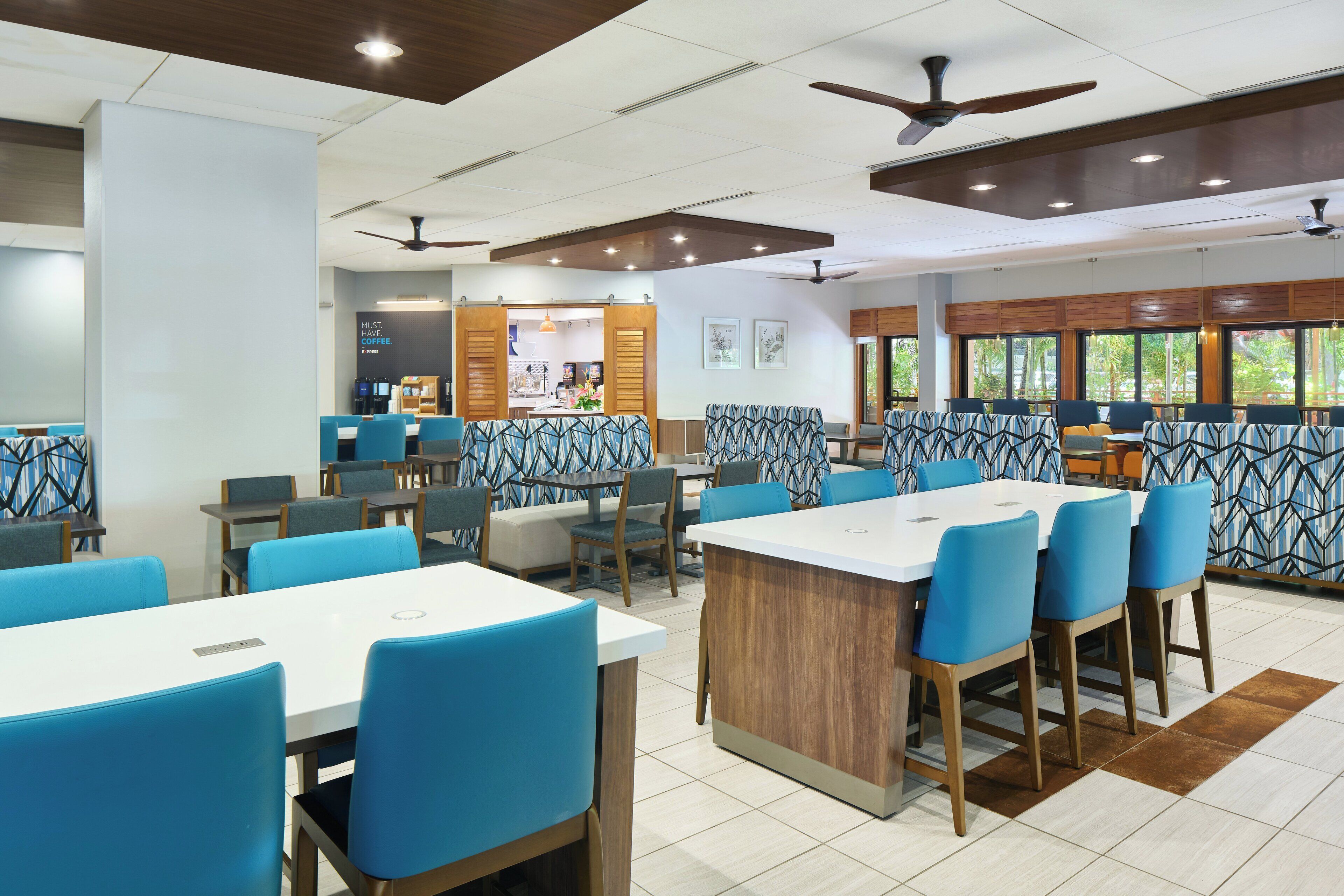 A modern dining area with high tables and blue chairs, a coffee station, and large windows with a view of greenery.