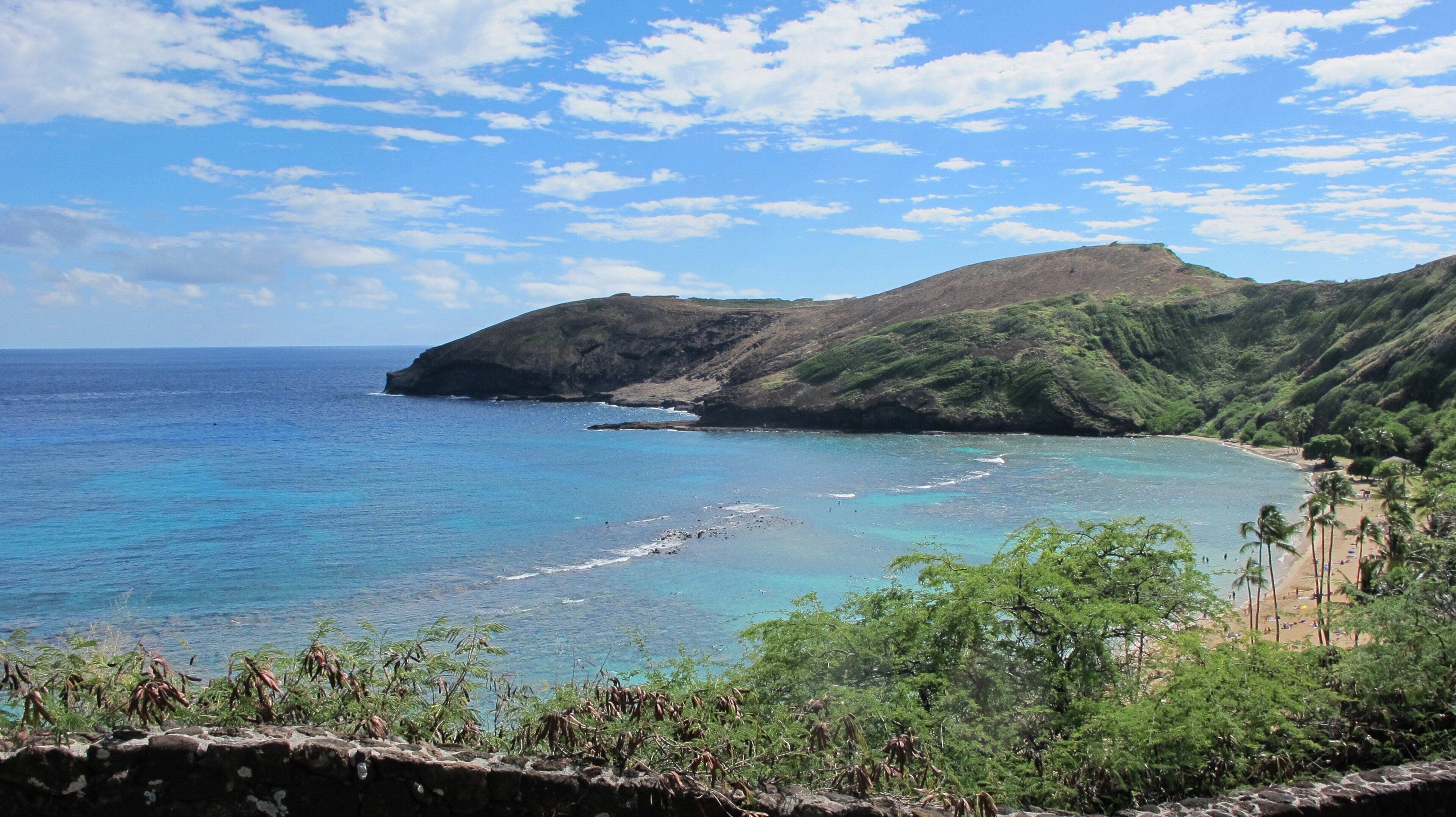 A coastal landscape with a beach, clear blue waters, and a mountainous backdrop.
