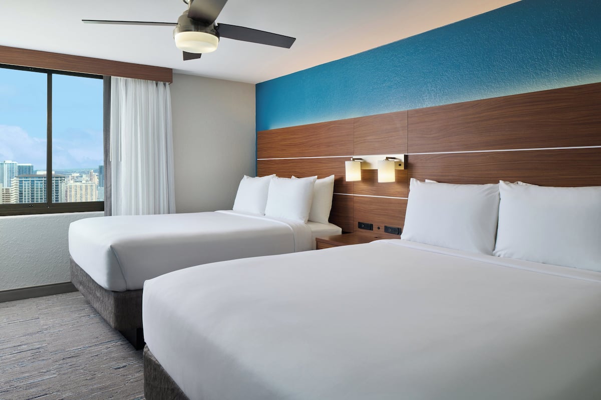 A hotel room with two beds, a large window with a city view, a ceiling fan, and a blue accent wall.