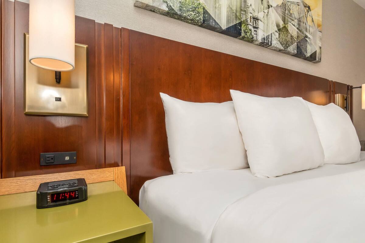 A hotel room with a wooden headboard, a bedside table with a clock, and a wall-mounted lamp.