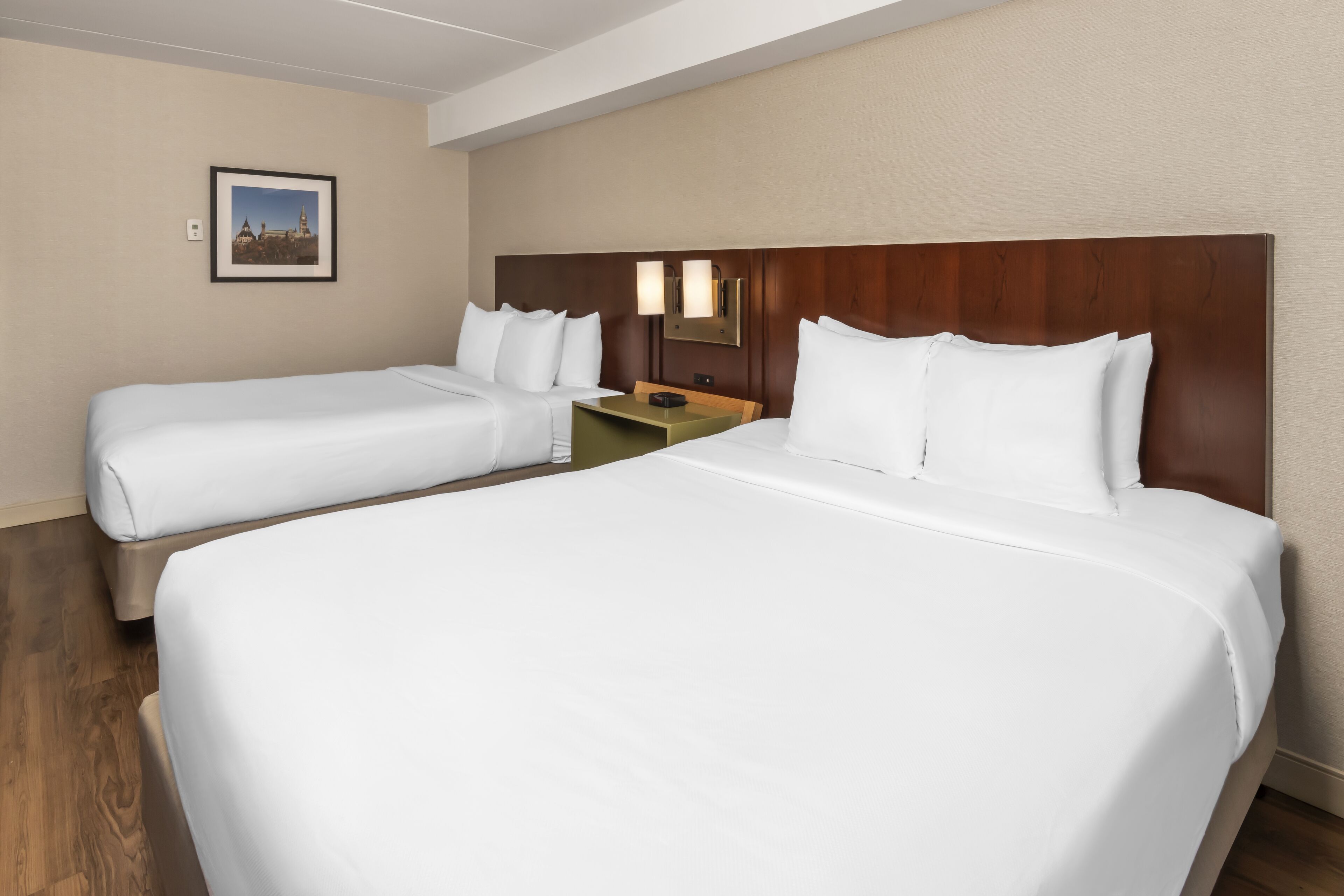 A hotel room with two beds, a wooden headboard, a nightstand, and a framed picture on the wall.