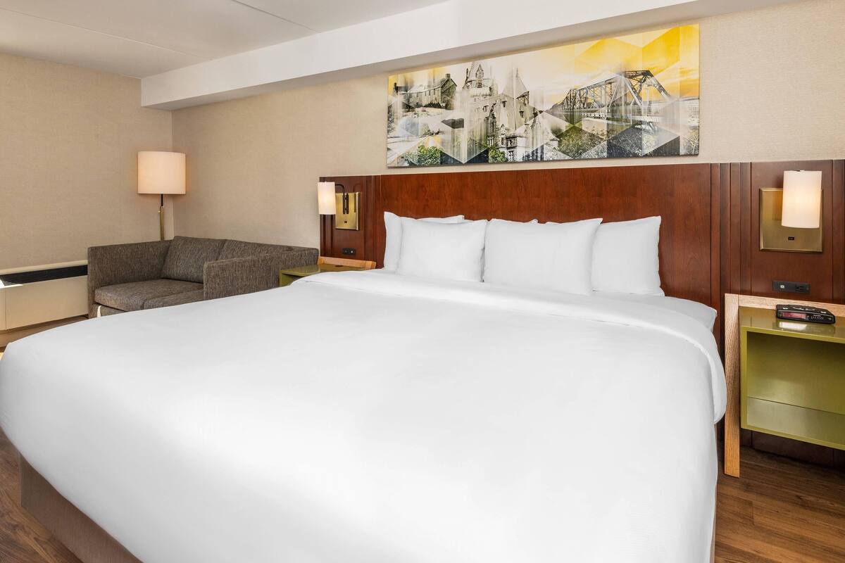 A hotel room with a large bed, a sofa, a nightstand with a lamp, and a painting on the wall.