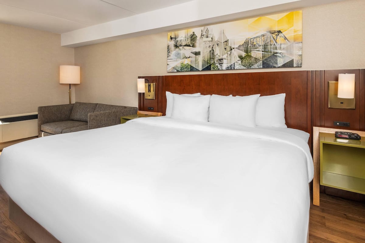 A hotel room with a large bed, a sofa, a nightstand with a lamp, and a painting on the wall.