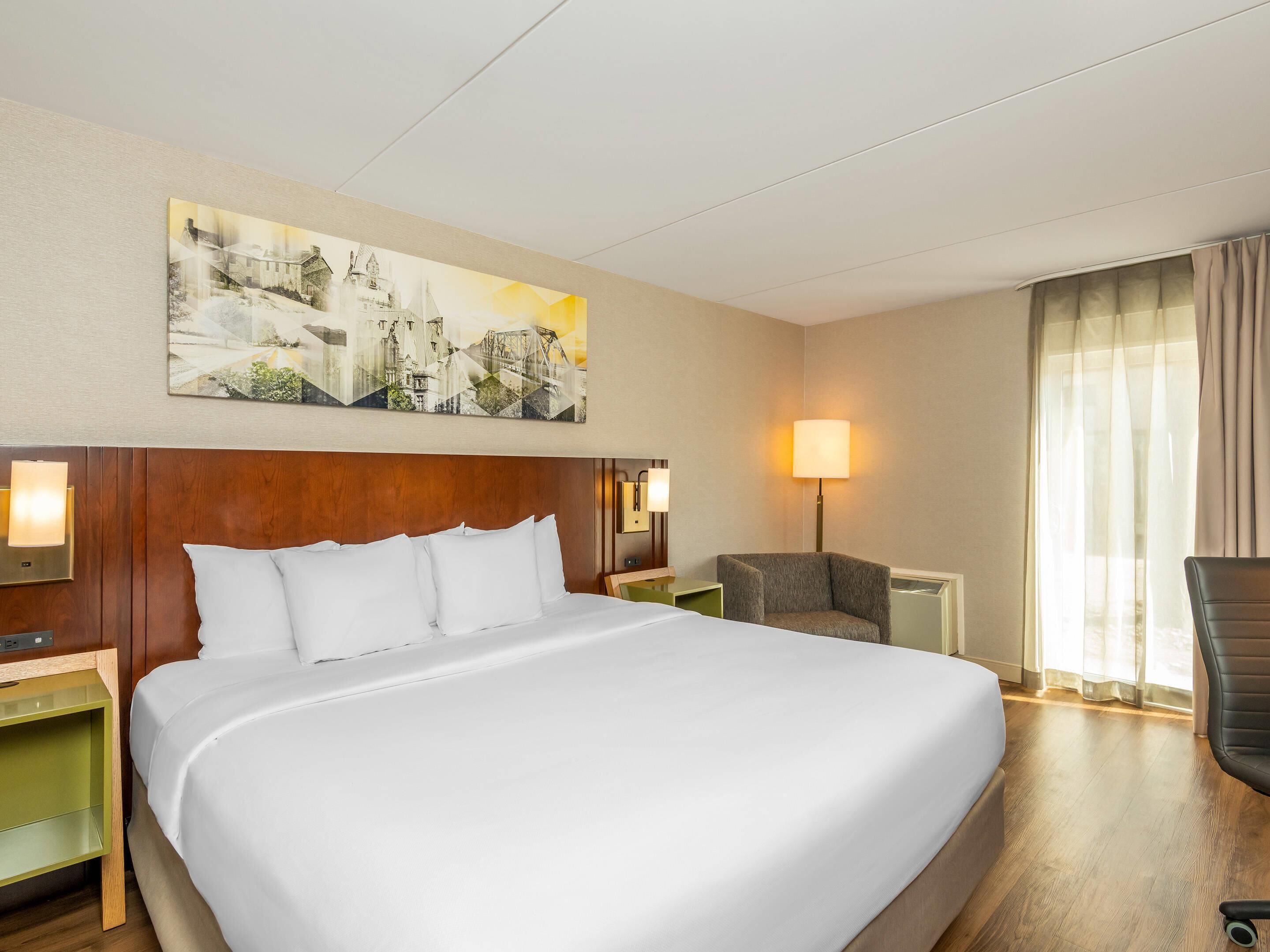 A hotel room with a large bed, a chair, a desk, a lamp, and a painting on the wall.