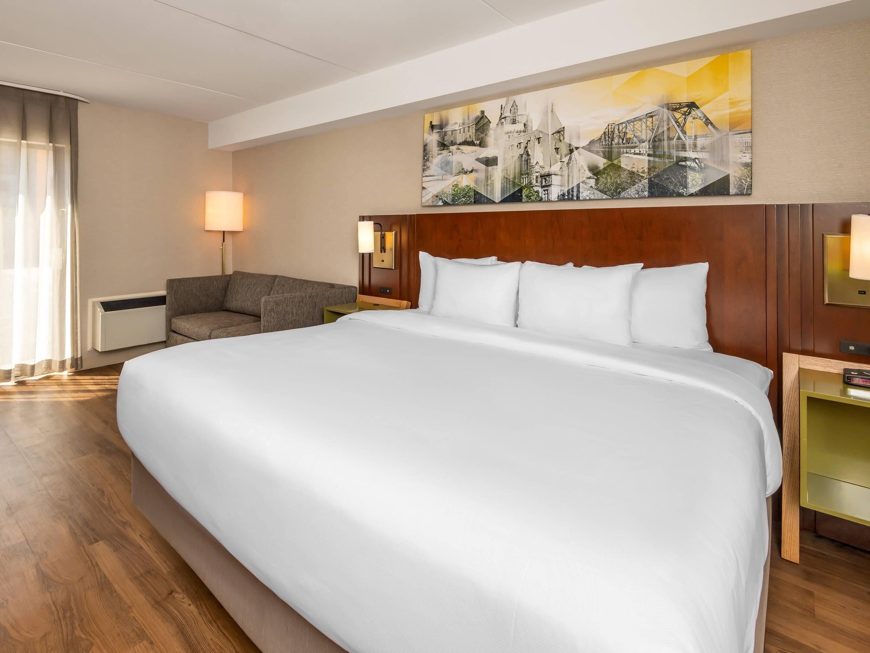 A hotel room with a large bed, a wooden headboard, a desk with a lamp, a chair, and a painting on the wall.