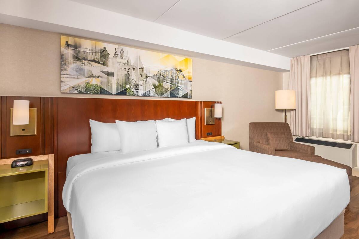 A hotel room with a large bed, a wooden headboard, a nightstand with a lamp, a chair, and a painting on the wall.