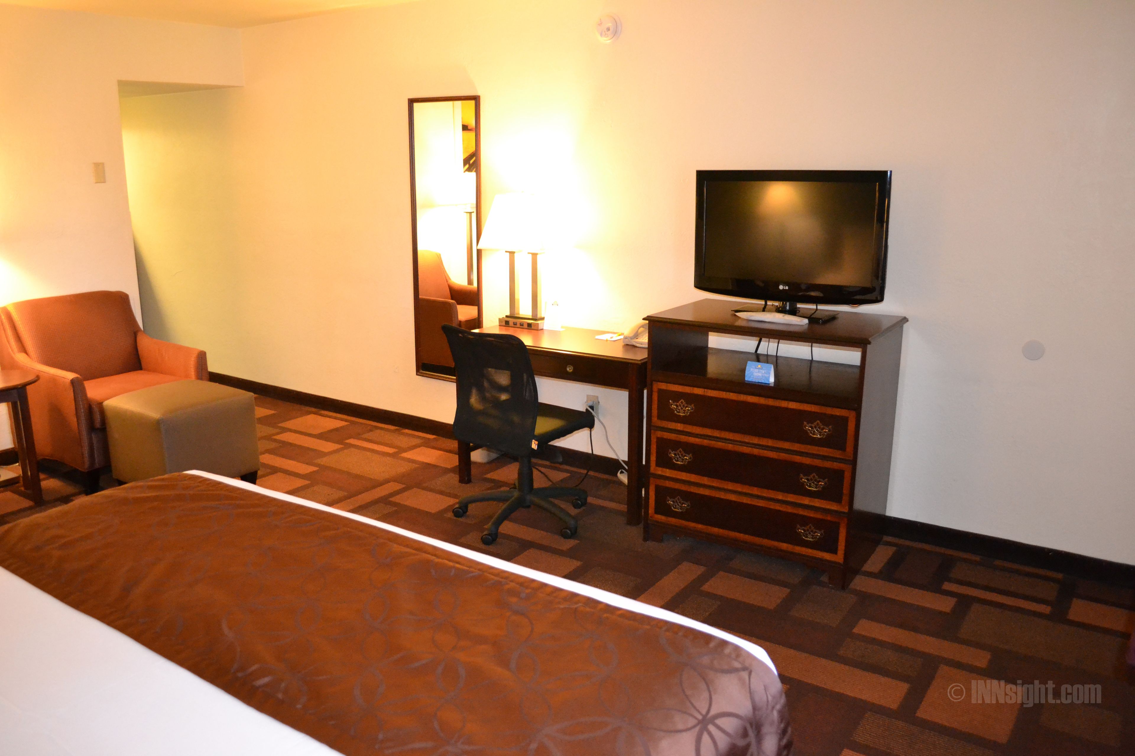Quarto de hotel com cama, escrivaninha com computador, cadeira, televisão e cômoda.