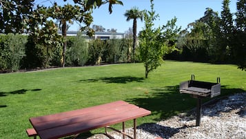 Una mesa de picnic y una parrilla en un jardín bien cuidado, con césped y varios árboles.
