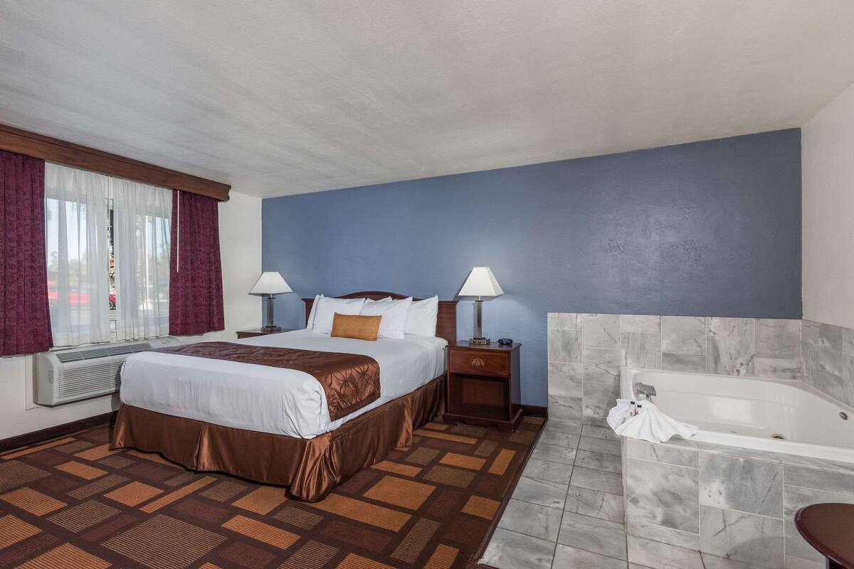 A hotel room with a large bed, two bedside tables with lamps, a window with curtains, an air conditioner, a bathroom with a bathtub, and a patterned floor.